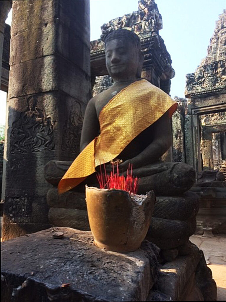 A stone male statue sits cross-legged with a golden fabric sash draped across his shoulder and chest and with of golden bowl filled with red incense sticks in front of him within the ancient temple of Angkor Wat in Cambodia.