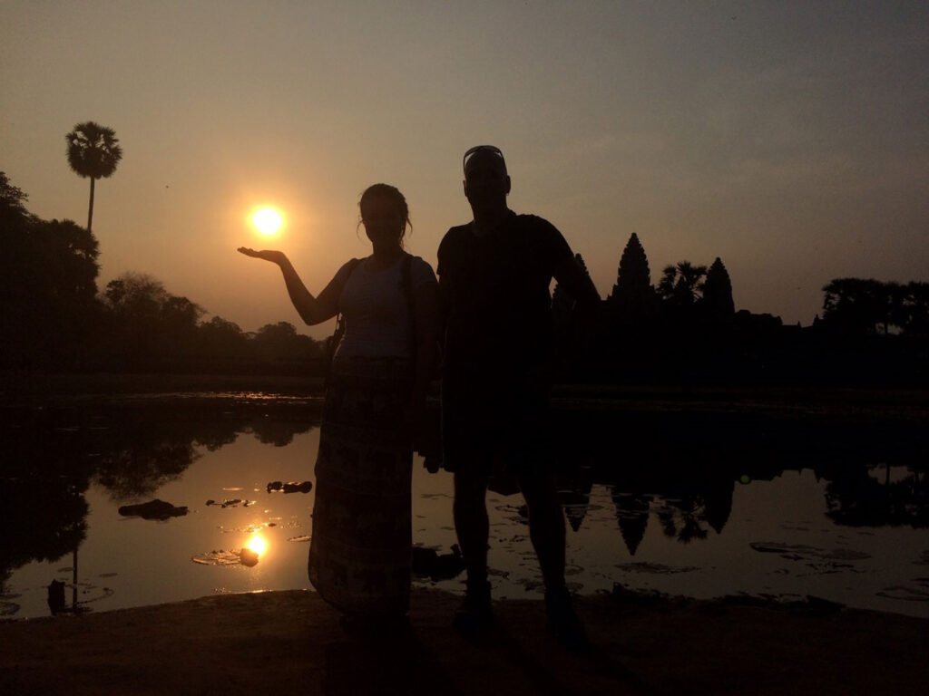 A man (pictured right) and a young woman (left in a long skirt) pose in semi-silhouette in front of a pond at sunrise outside Angkor Wat temple in Cambodia with the woman lifting her right arm upward, palm open, as if to be holding up the rising sun.