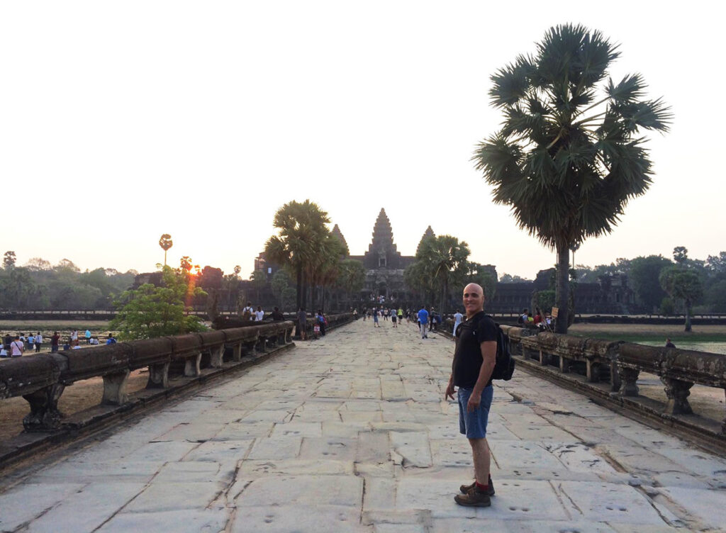 Inner Space host, Ralph Williams, stands at sunrise on a wide stone bridge-pathway that is lined with palm trees, leading to the large Cambodian temple, Angkor Wat.