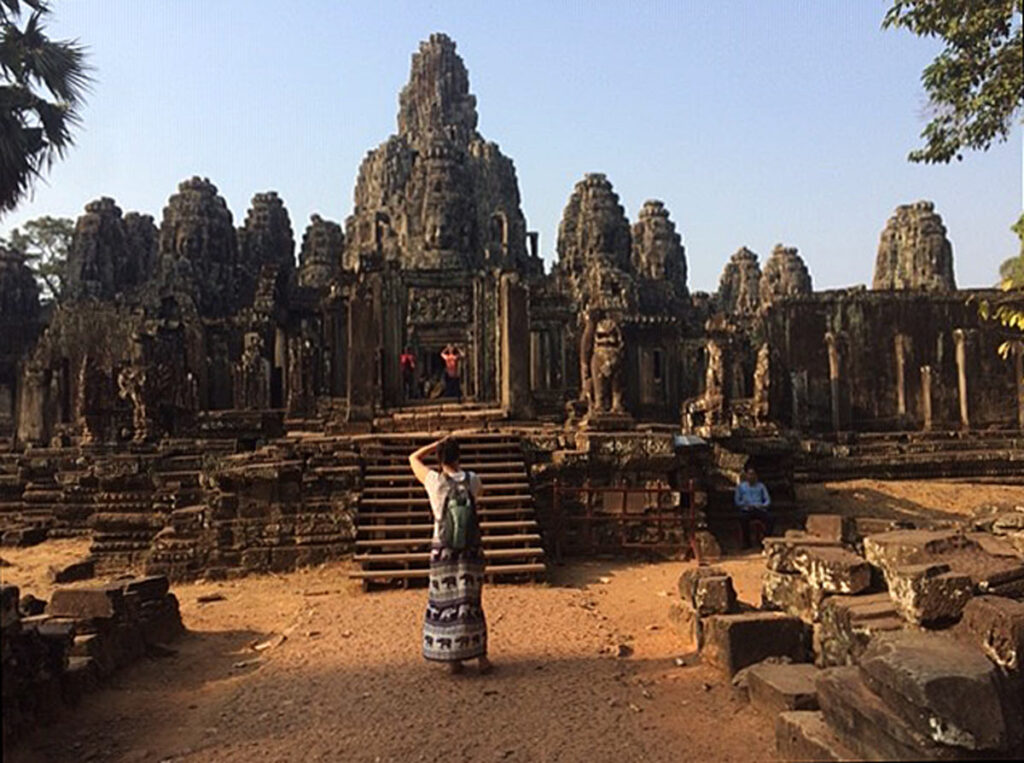 A woman in a long skirt stands in front of and photographs an ornate temple-relic made of stone.