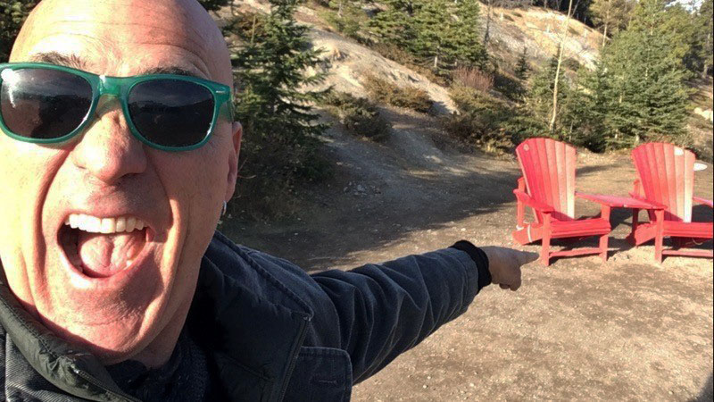 Person with green sunglasses pointing to two red Adirondack chairs in an outdoor scenic setting.