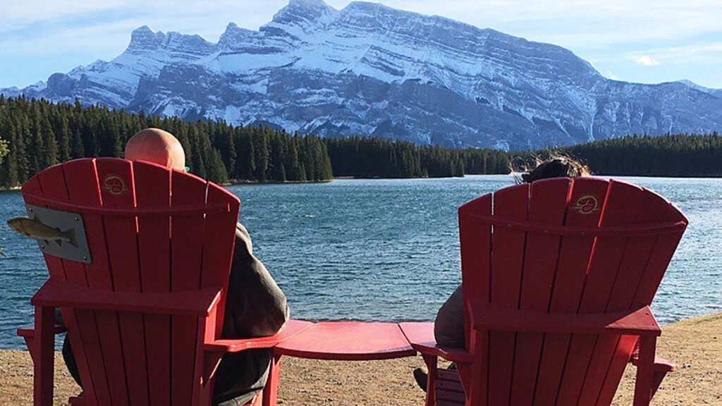 Inner Space host, Ralph Williams, and his daughter sit in a pair of red Adirondack chairs that face a small tree-lined lake with snowy mountains in the background in Banff National Park in Canada.