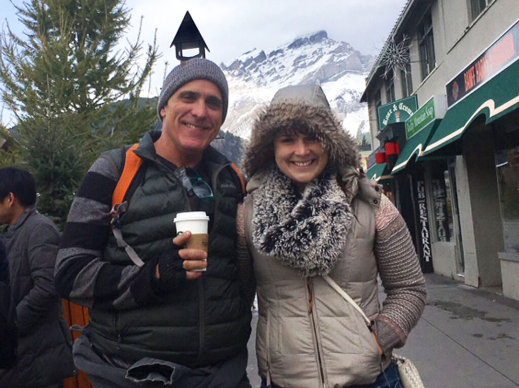 Inner Space host, Ralph Williams (dressed in winter clothing and holding a paper coffee cup) poses for a photo with his daughter, Phoebe, along the streets of Banff, Canada, with a snowy mountaintop in the background.