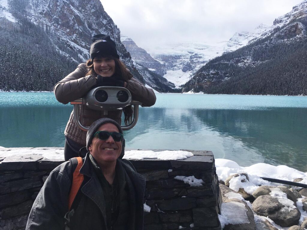 Gorgeous view of the clear turquoise waters of Lake Louise and the surrounding snow-capped mountains with Inner Space host, Ralph Williams, on the ground and daughter Phoebe leaning on a binocular-viewer, smiling.