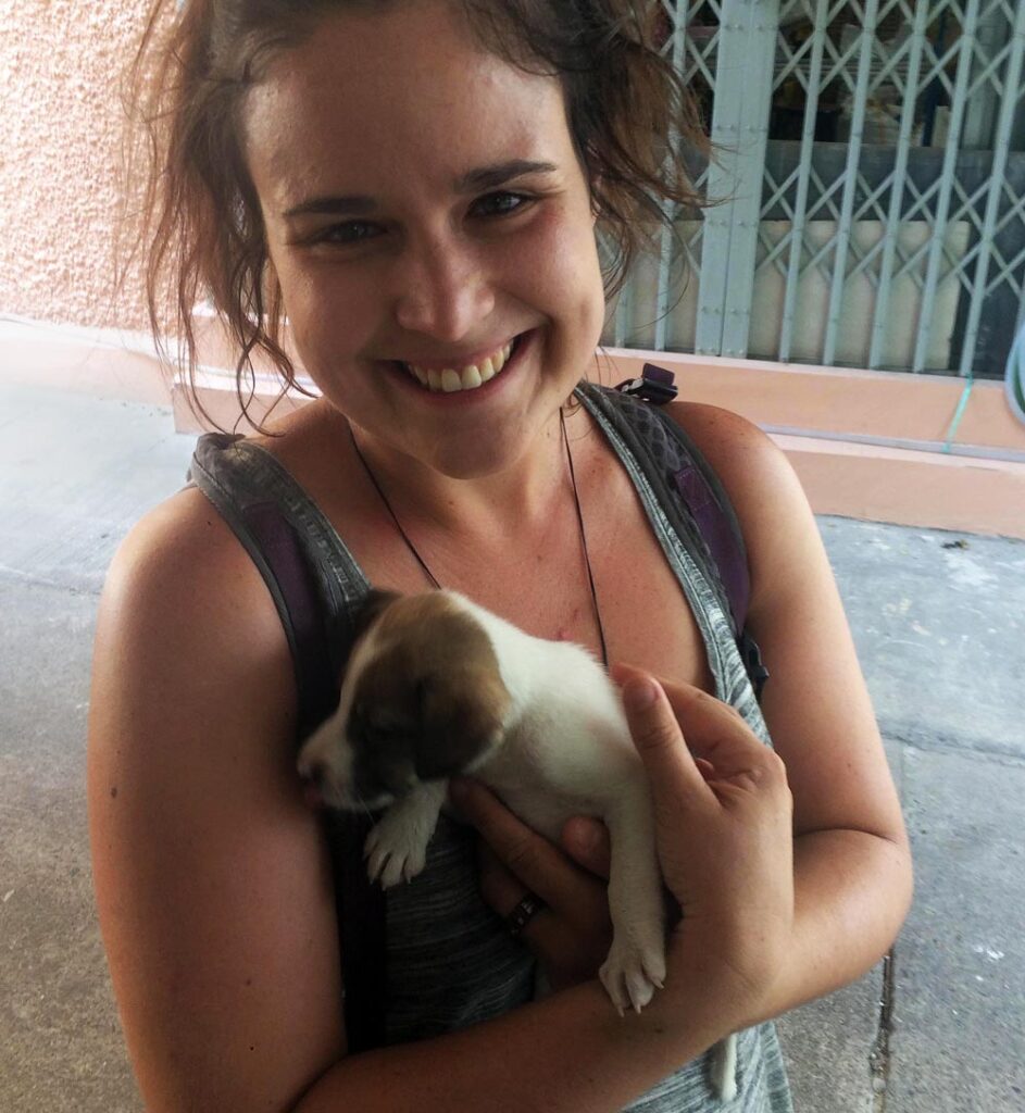 Person outdoors, smiling and holding a small puppy, with urban gated structure background.