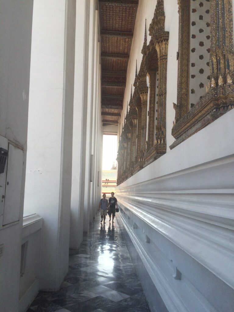 Two peope in semi-sihlouette walk down a narrow walkway with white columns on one side and intricate gold frames on the other in a temple in Bangkok, Thailand.