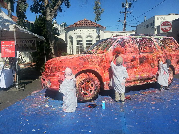 Children painting a colorful SUV under a tent outdoors with a building labeled "Athenaeum" in background.