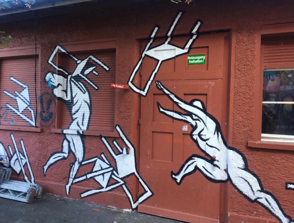 Street art mural with white, cubist figures on a reddish-brown wall, windows, door, and green sign.