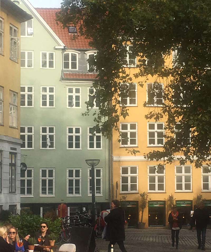 Colorful urban scene with pastel green and yellow buildings, white windows, outdoor café, and trees.