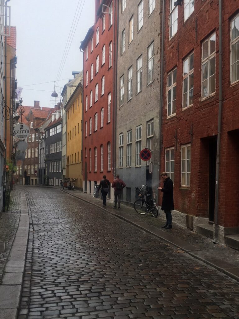 View of a narrow cobbled street lined by colorful brick buidlings with a young man in a black coat smoking a cigarette on a damp morning in Copenhagn, Denmark.