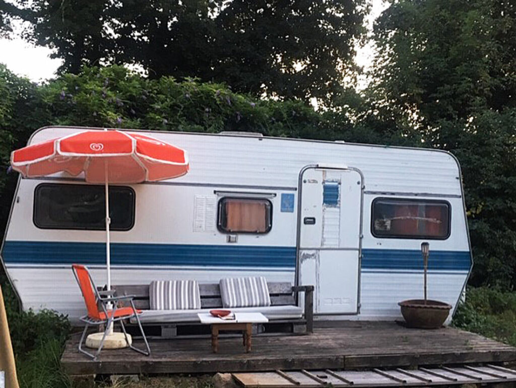 Vintage white and blue camper trailer with a wooden deck, seating area, and patio umbrella in greenery.