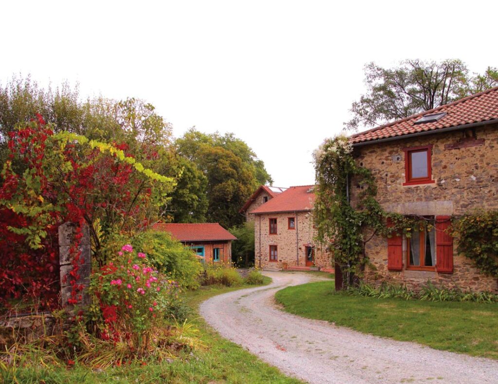 Stone houses with red roofs and shutters, surrounded by greenery, flowers, and a curved gravel path.