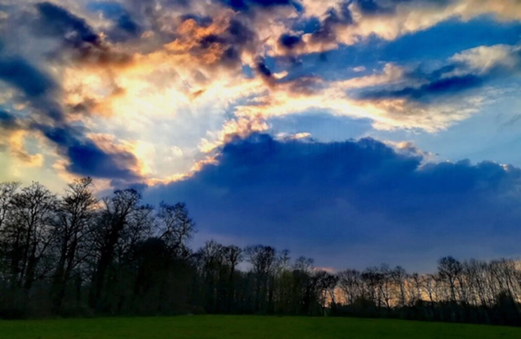 Sunlight bursts through the late-afternoon clouds over trees lining bright green meadow in Limousin, France.