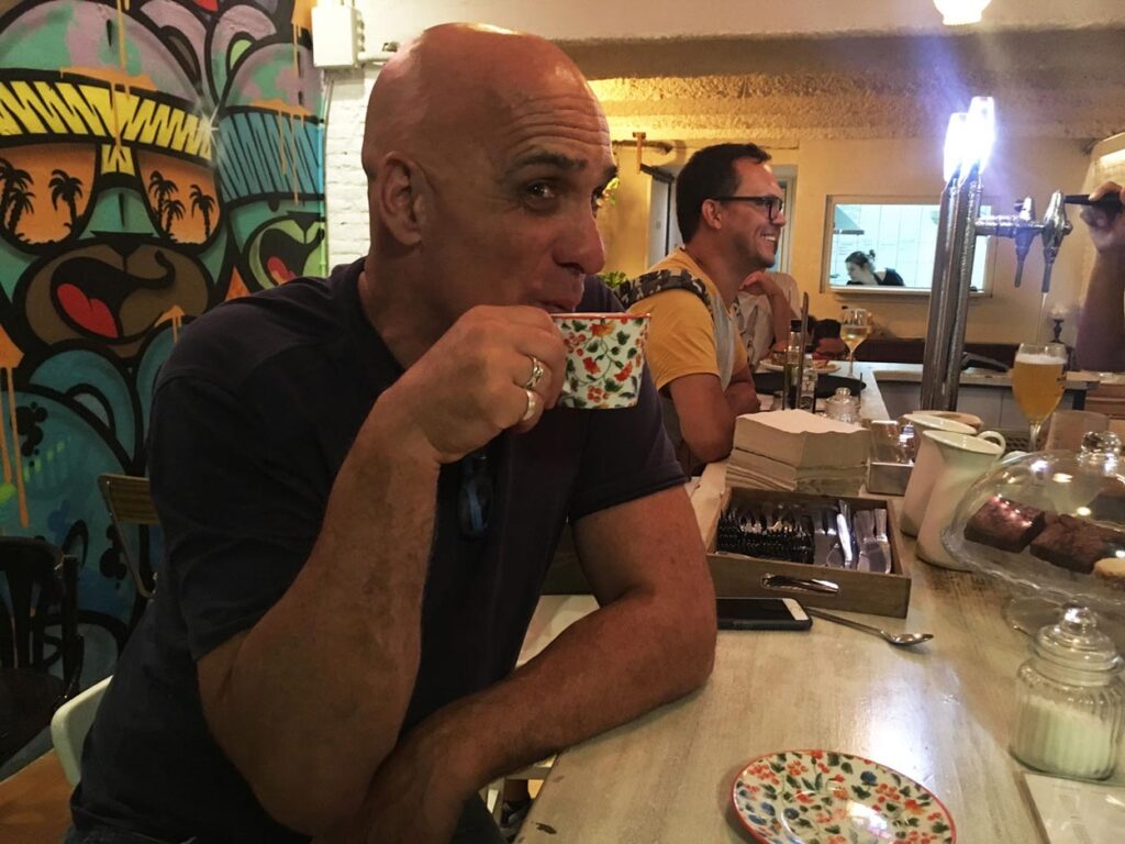 Two men at a bar counter with tropical mural, sipping drinks, pastries and utensils on the counter.