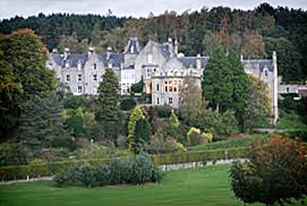 Large historical stone building with gables and chimneys, surrounded by lush gardens and dense trees.