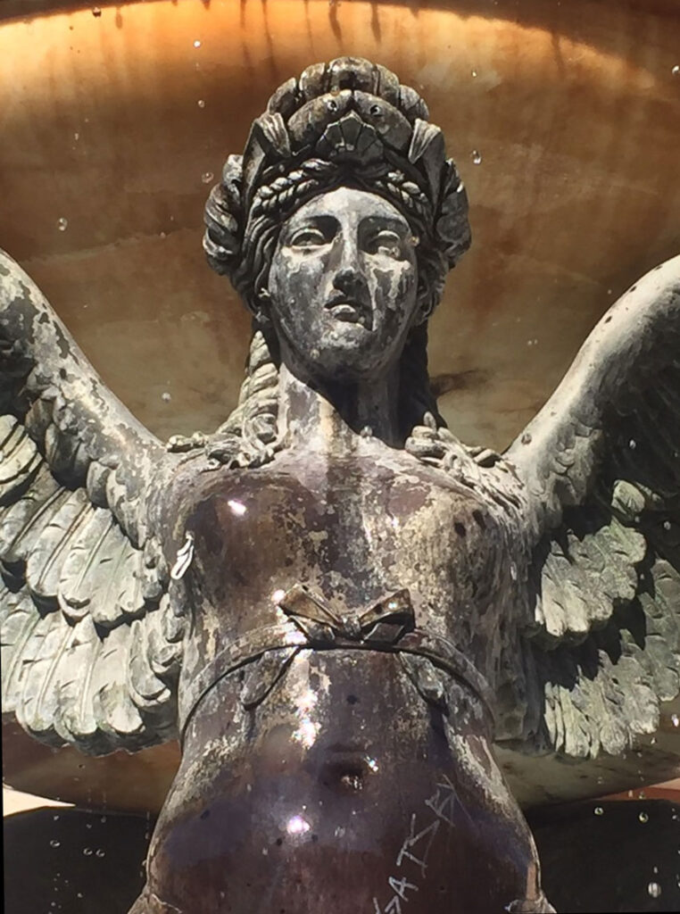 Weathered winged female statue with intricate headwear in a fountain setting.