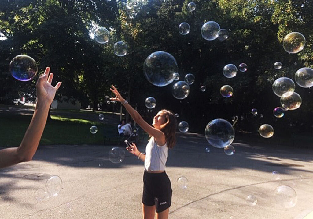 A teenage girl reaches upward toward a collection of large soap bubbles drifting by in a sunlit park in Zurich, Switzerland.