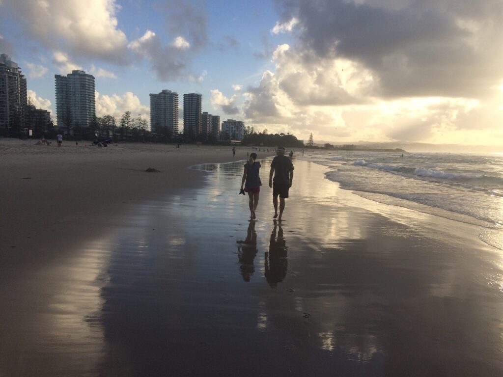 A couple in silhouette walks barefoot in the wet-packed, reflective sand near the water's edge near-dusk with highrise buildings in the background.