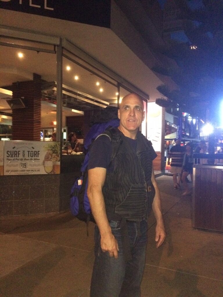 Person with dark vest and blue backpack outside an illuminated restaurant with "Surf & Turf" signage.