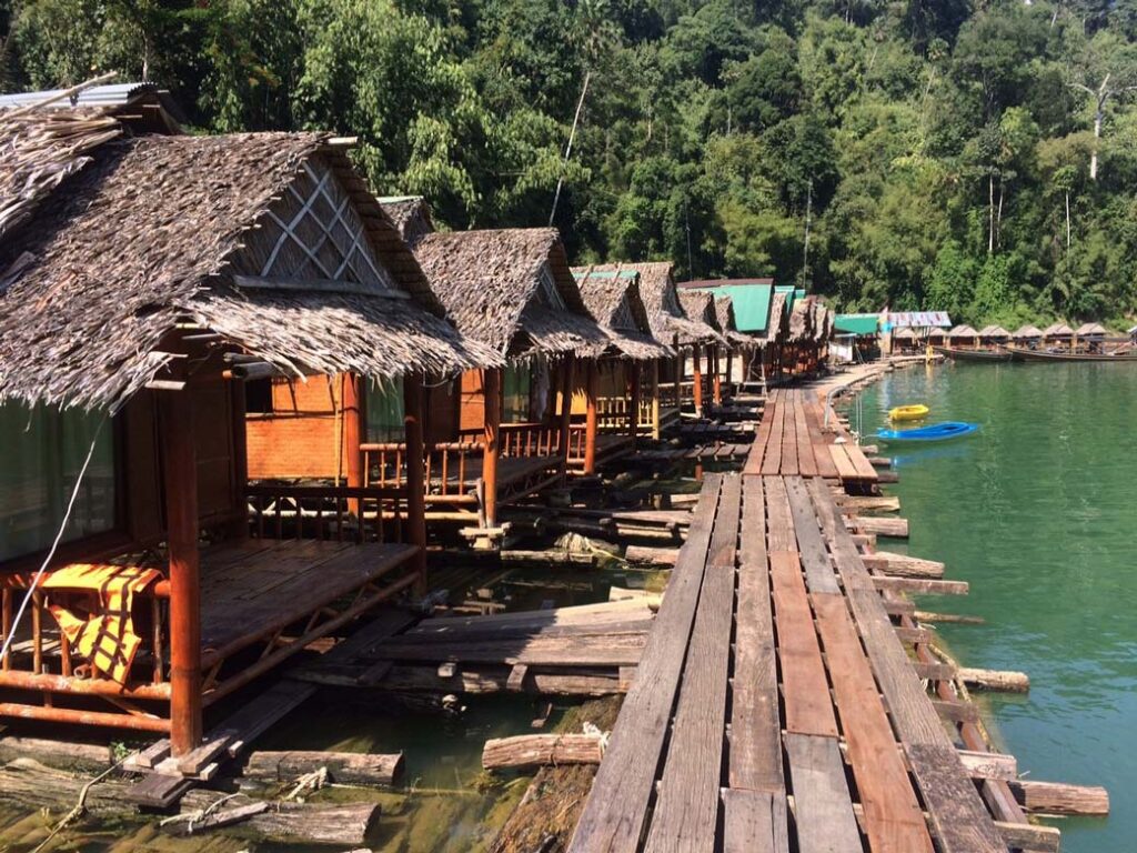 A long row of vacation huts featuring thick grass roofs and wooden front porches sit directly on a body of water with a wood-planked walkway in front and a steep hill of green trees in the background.