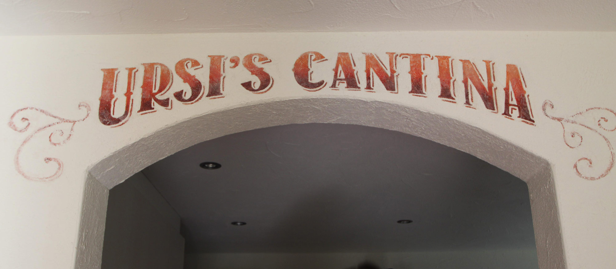 Portfolio photo of some hand-painted western-style lettering that reads, "URSI'S CANTINA" in sunset orange( on white walls) that Inner Space host, Ralph Williams, painted above an arched doorway to a kitchen for a client in Switzerland.