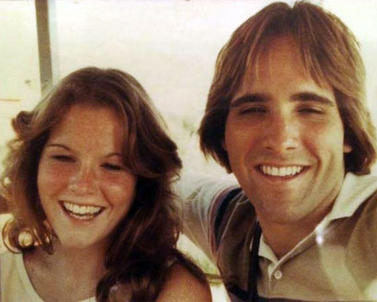 Young man and woman smiling outdoors, man with shoulder-length brown hair, woman with wavy brown hair.