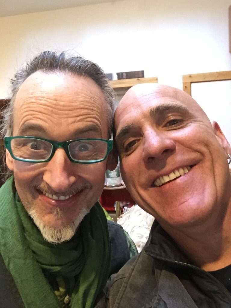 Two smiling individuals close together; one wears green glasses and scarf, the other has a bald head and earring.