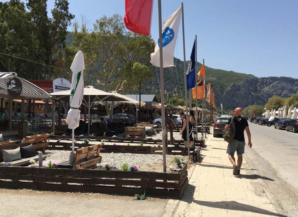 Pedestrian walkway with flags, outdoor seating, boutiques, parked cars, mountains, and trees.