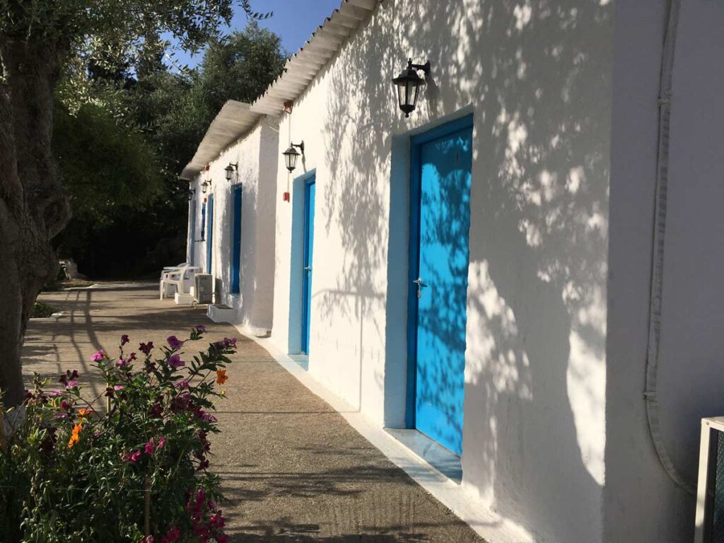 Row of white buildings with blue doors, lanterns, tree shadows, and colorful flowers on pathway.