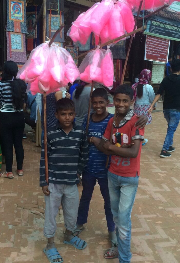 Three smiley-faced Nepali boys stand together, posing for the photo, while holding long wooden sticks with a hot-pink substance (cotton-candy?) at the top, for sale in plastic bags.