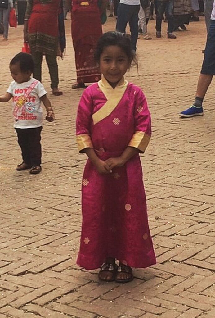 A young child in a traditional pink and gold outfit stands on a stone-paved area, with people in the background.