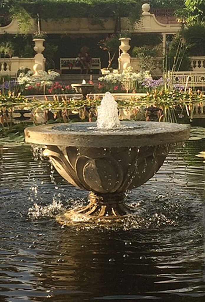 Stone fountain with a tiered design in an ornamental pond, surrounded by lush greenery and flowers.