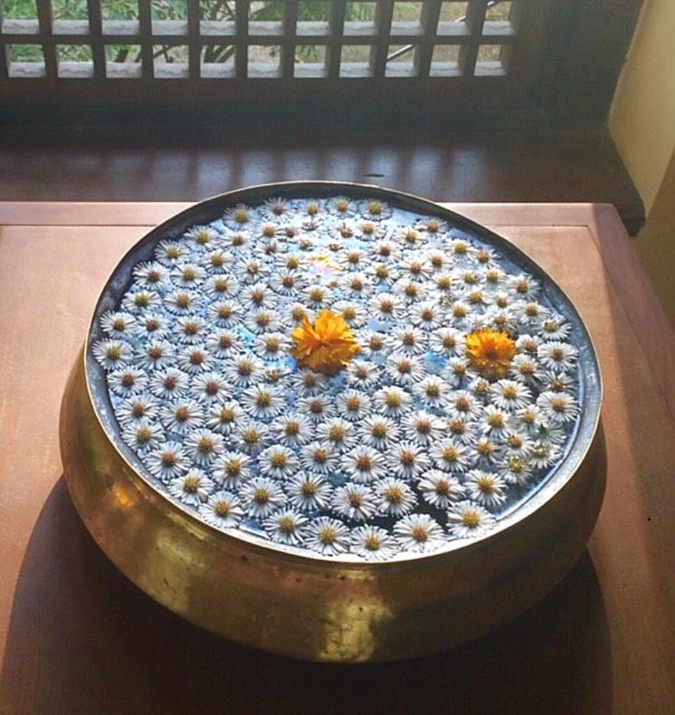 Tiny white flowers and two yellow blossoms float in a gold bowl on a small wooden table with sunlight streaming in through a grid-like wooden window within the "Garden of Dreams" Botanical Park in Kathmandu.