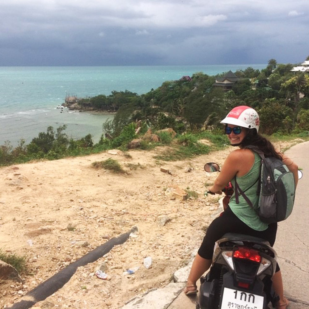 Person on scooter with helmet and sunglasses overlooking coastal view with greenery and ocean.