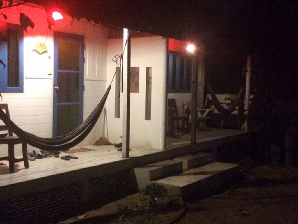 Porch of a small building at night with red light, hammocks, chairs, and broom near a blue door.