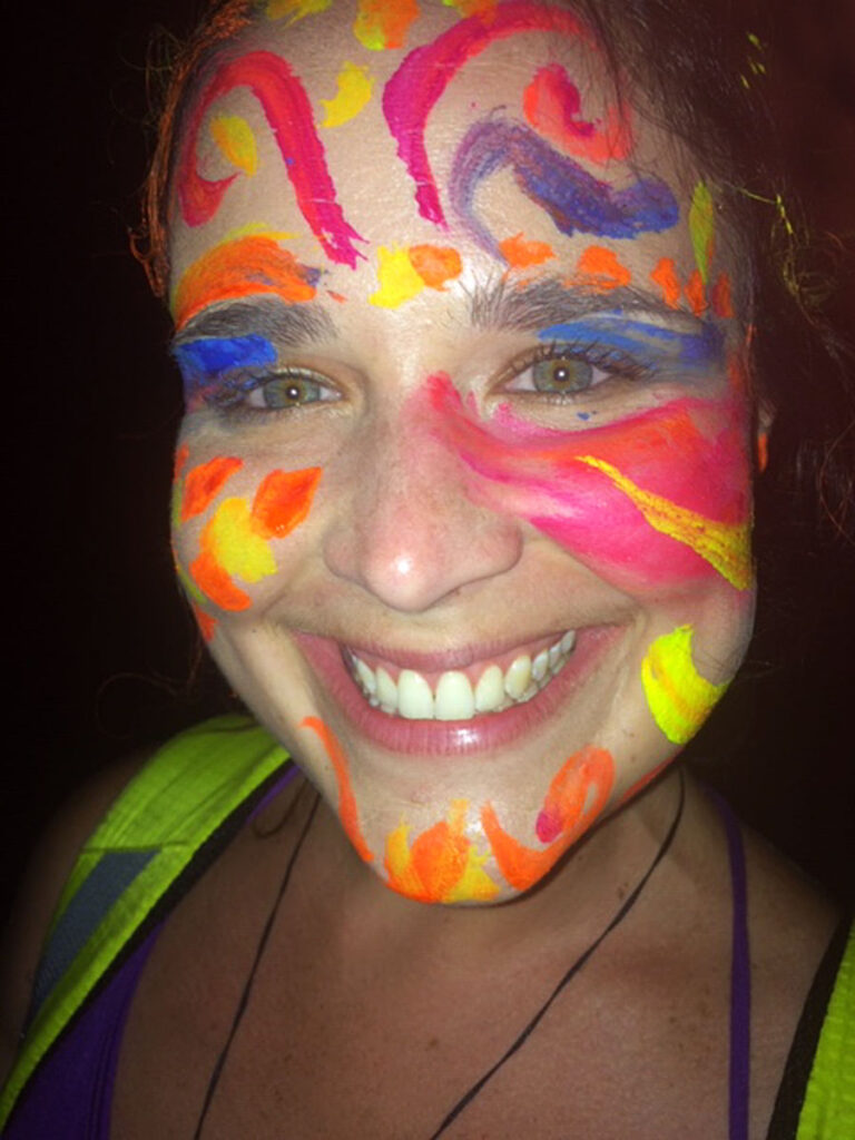 Smiling person with vibrant neon face paint in abstract patterns, pictured in a dark environment.