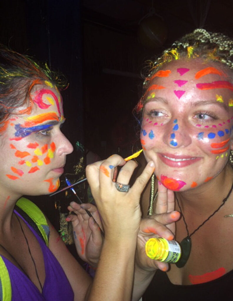 Face painting activity with vibrant neon colors, one person applying paint on another at a festive event.