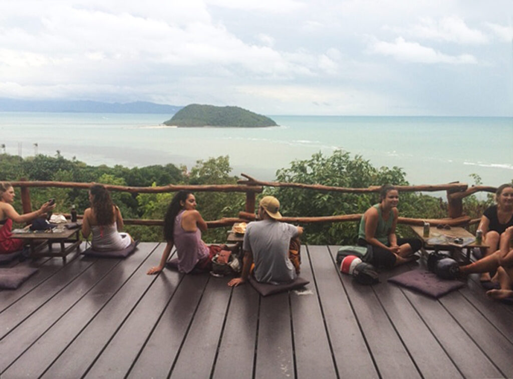 Group of people seated on an outdoor wooden terrace enjoying an ocean view with a small island.