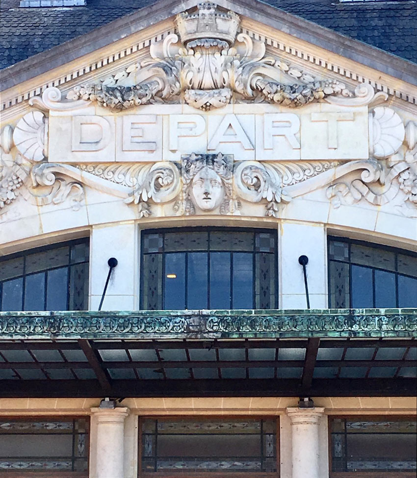 Building facade with "DEPART" inscription, ornate carvings, central crown, face motif, arched windows.