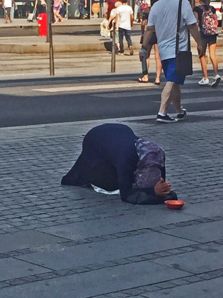 Person in dark clothes kneeling with headscarf, orange bowl on pavement, pedestrians in background.