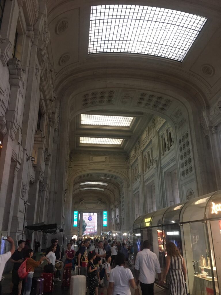 Busy train station interior with high arched ceilings, skylights, people, luggage, and retail shops.