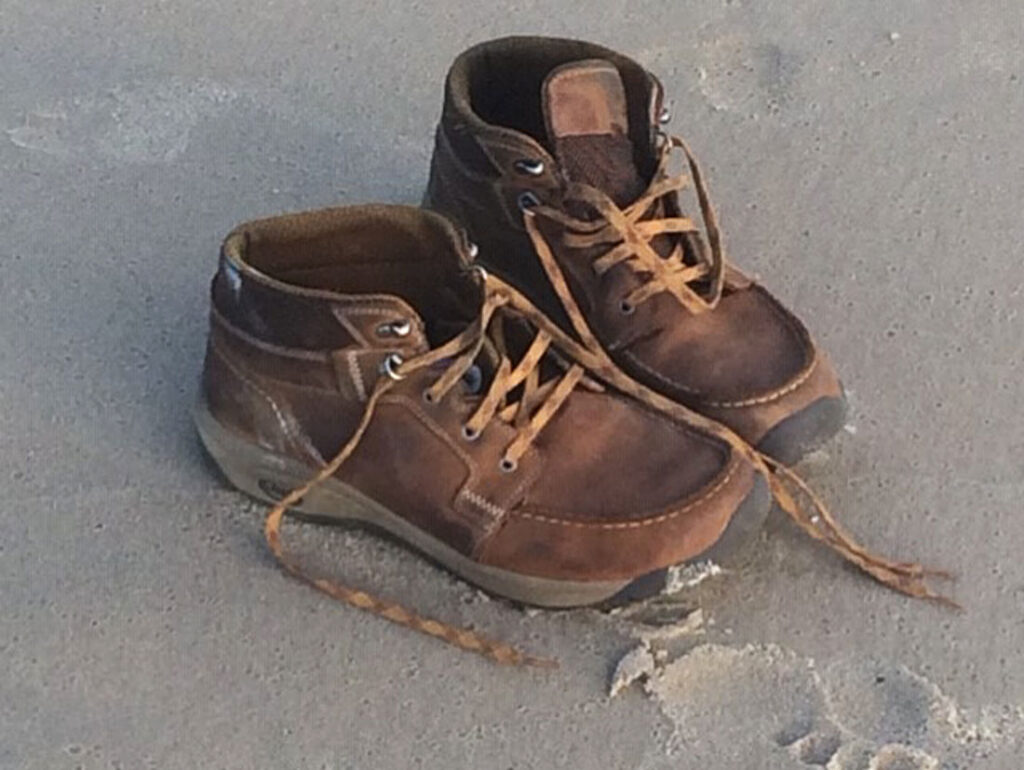 Worn brown leather boots with untied laces on a sandy surface.