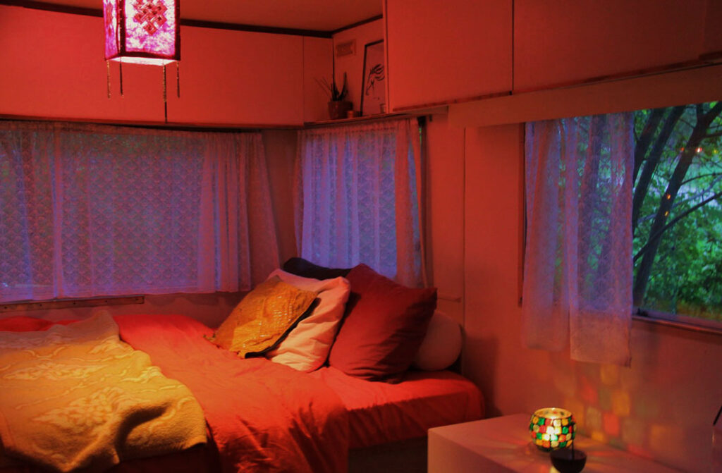 Cozy bedroom with warm lighting, a bed, lace curtains, lantern, colorful lamp, and green tree view.