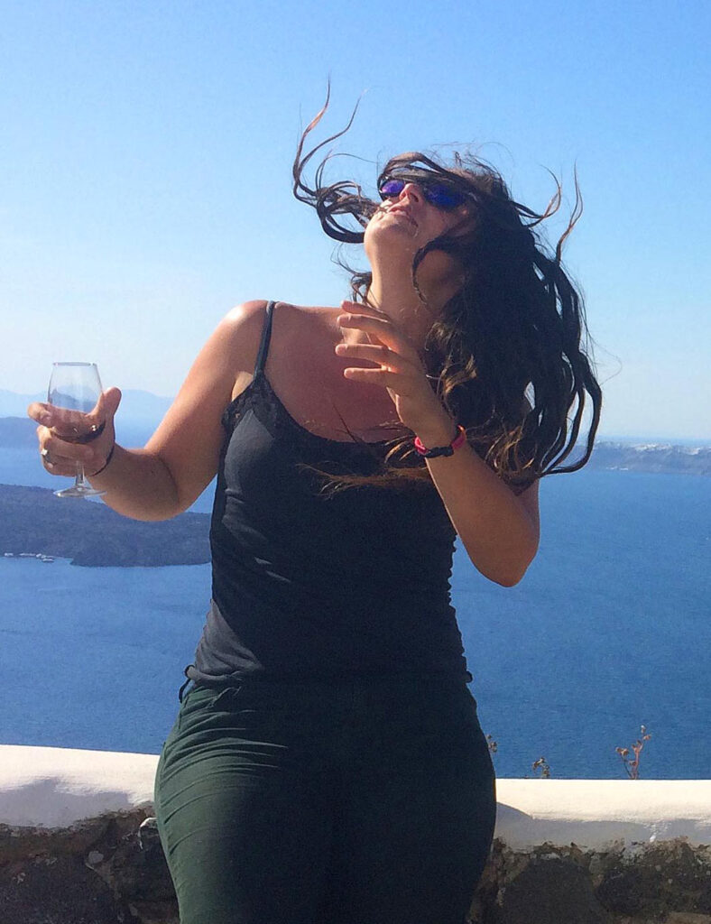 A young woman in a black dress sits on a low stone wall while holding a wine glass and looks upward as her hair blows in the wind.