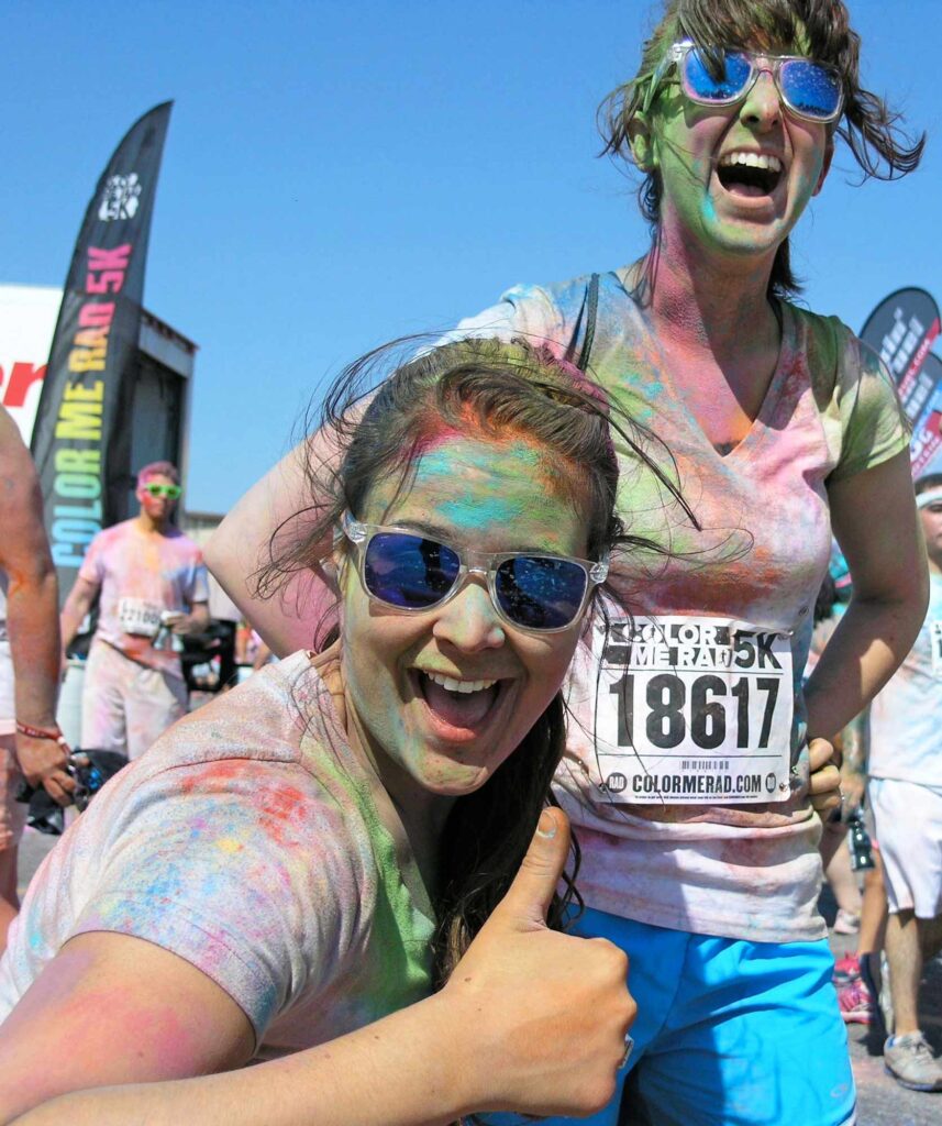 Two young women wearing sunglasses are covered in bold colors after completing a "Color Me Rad" 5k run in Oklahoma City (and getting dowsed with colors while en route), with one of them kneeling and giving a "thumbs up" while the other laughs from above.