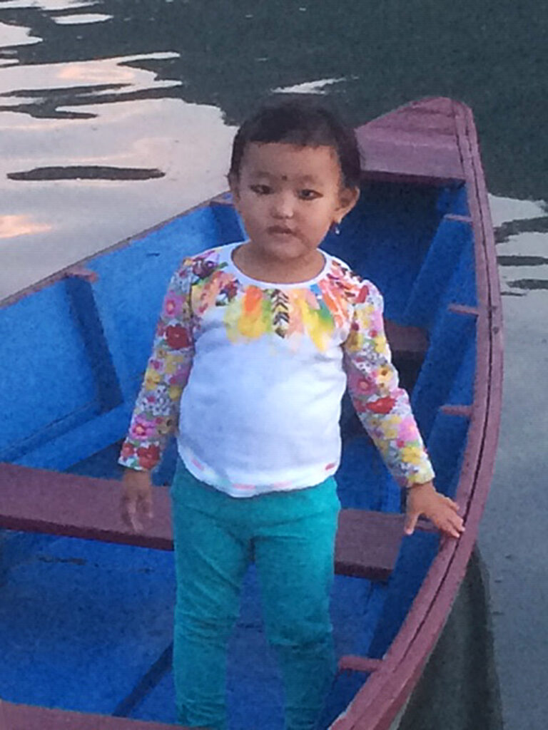 Young child in colorful shirt and green pants standing in a blue boat on calm water.