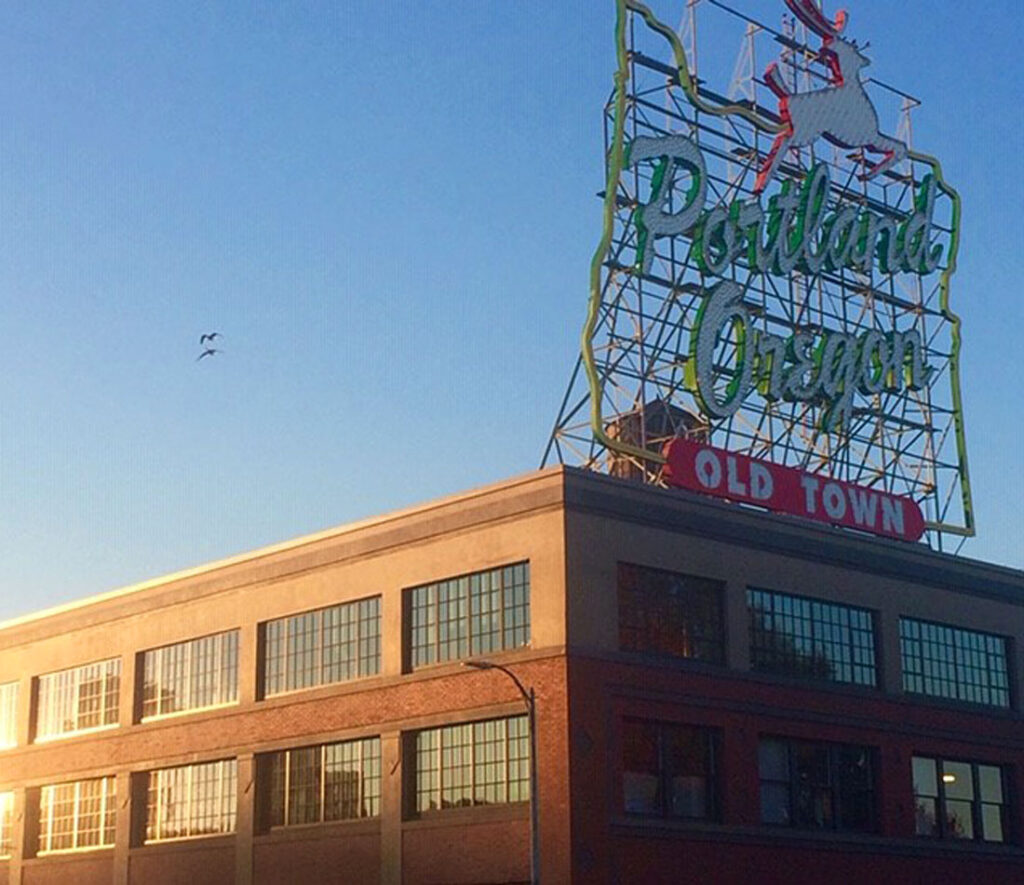 Building with large windows at sunset, "Portland Oregon Old Town" sign, deer silhouette, clear sky.