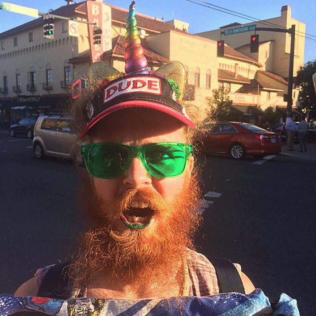 Funny headshot of a man with a scraggly red bead, bright green sunglasses, and a ballcap that says "DUDE" with a rainbow unicorn horn at the top.