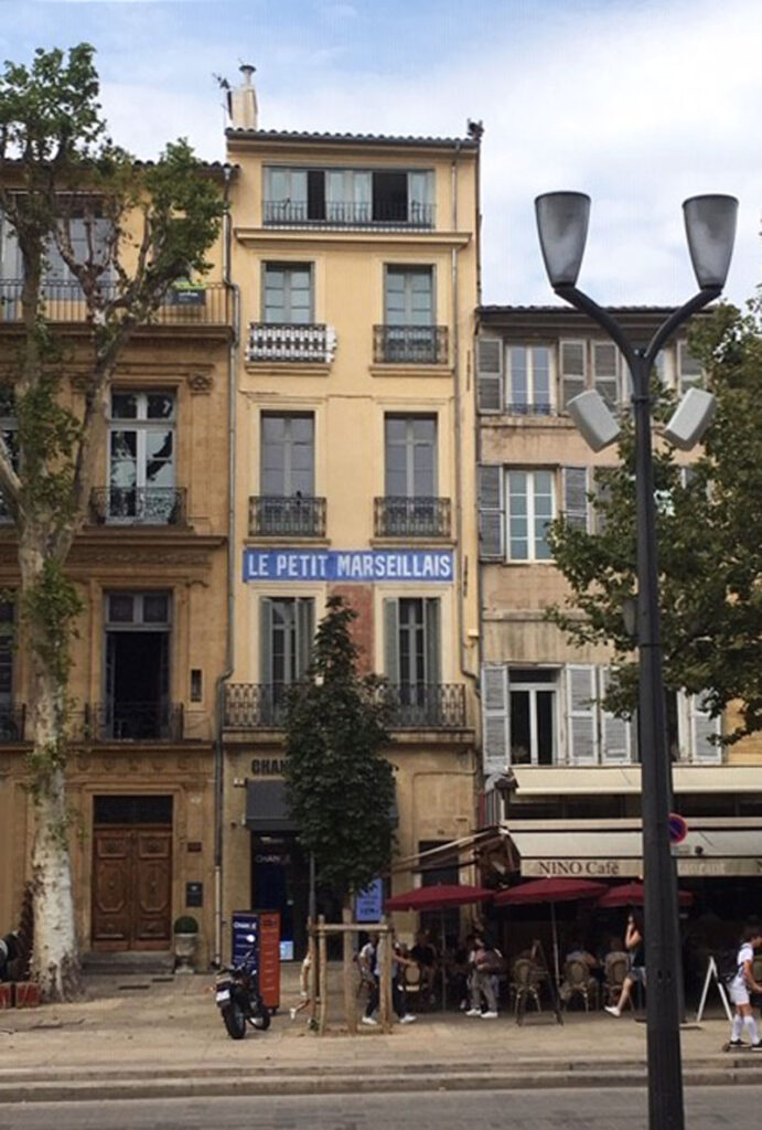 Multi-story yellow building with "Le Petit Marseillais" sign, trees, street lamp, and "Nino Café."