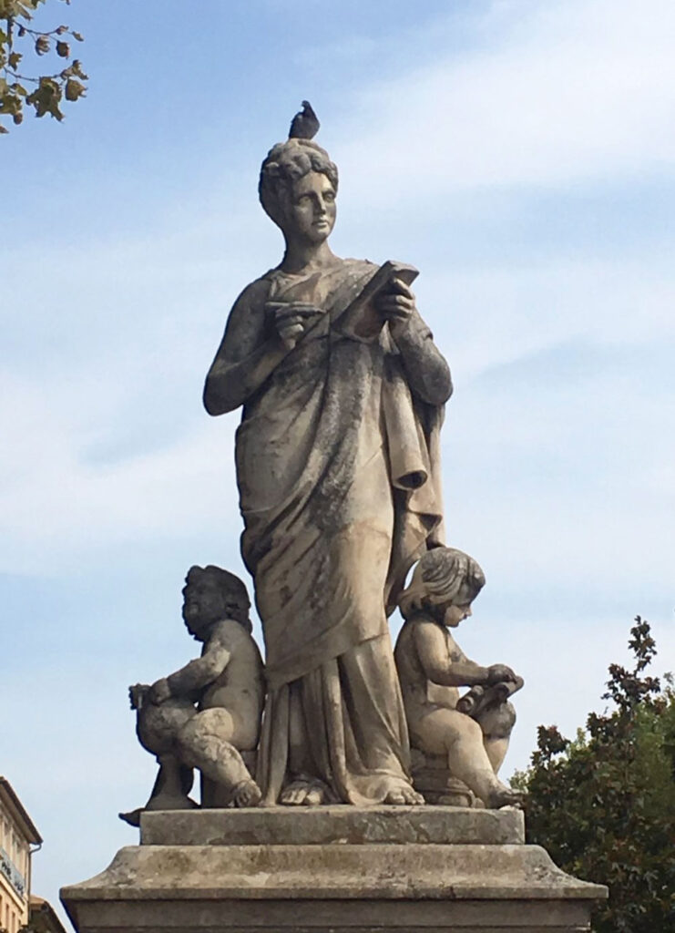 Stone statue of a robed woman with two children, pigeon on head, trees and buildings in background.